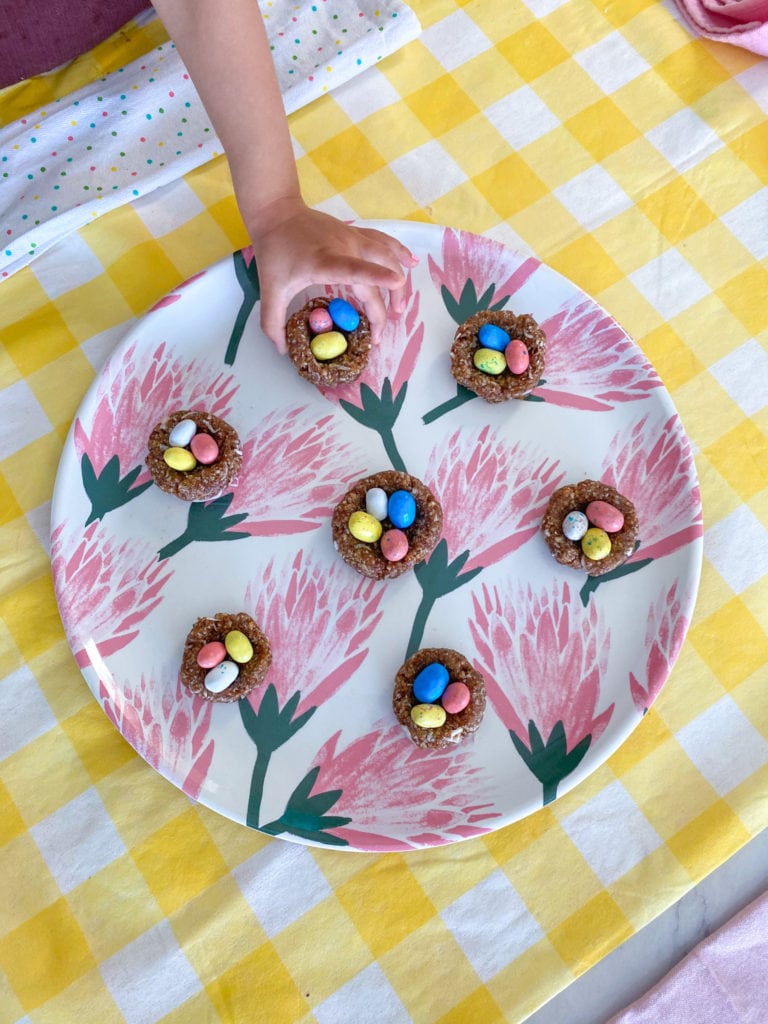 a toddler sized hand is trying to grab a colorful easter bird's nest treat from a plate full of them