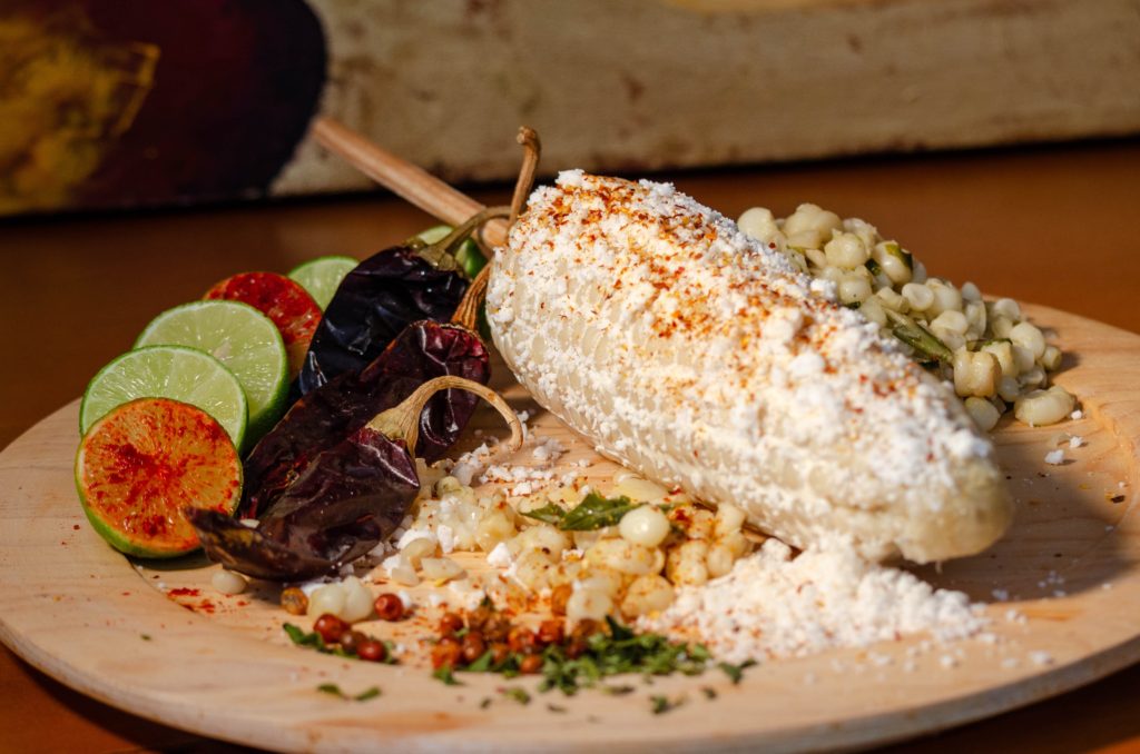 Elote served on a wooden plate with lime slices and chile.