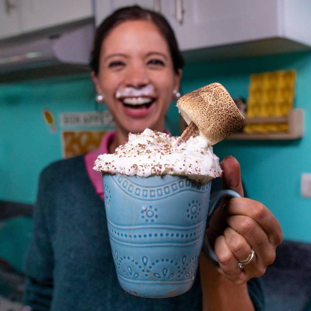 La Cooquette holding a huge mug of hot chocolate topped with whipped cream, a wafer, and a roasted marshmallow